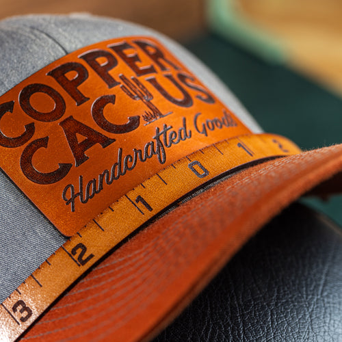 A close-up of a gray and orange hat with a patch on the front showing the text 'Copper Cactus' and a measuring tape indicating the size of the patch.