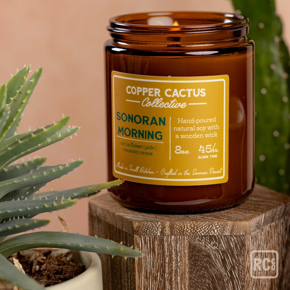 Candle jar labeled 'Copper Cactus Collective Sonoran Morning' on a wooden block with plants in the background.